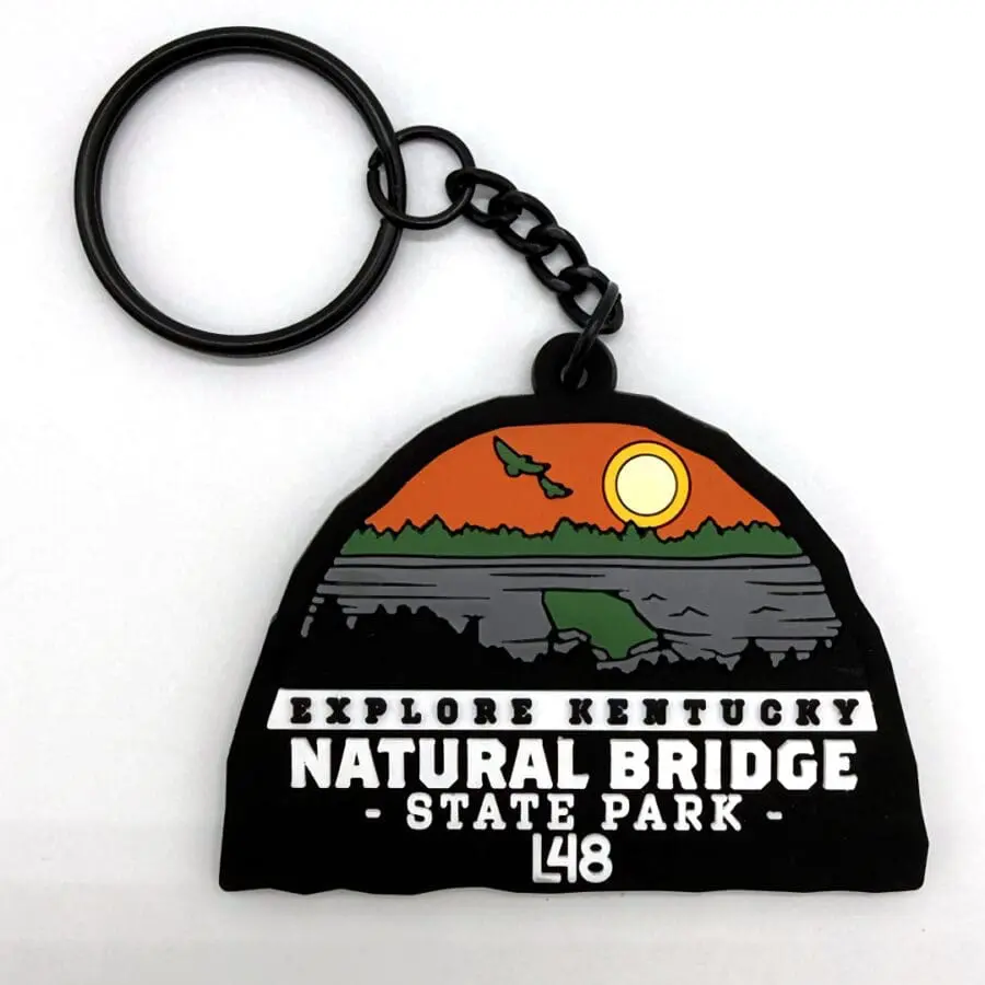 Souvenir Keychain Featuring a Stylized Sunset over a Landscape with Trees, Hills, and a Bridge, Text Reads 'explore Kentucky Natural Bridge State Park L48'